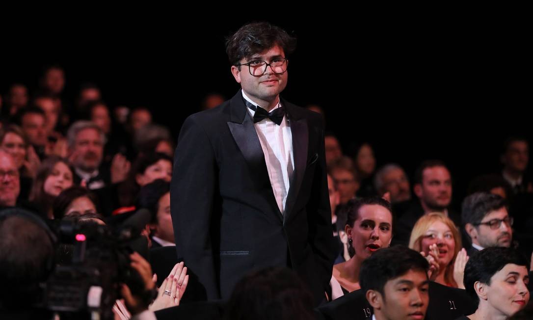 O diretor, um homem branco usando óculos e terno preto, está em pé em meio a uma plateia no Festival de Cannes, ele sorri