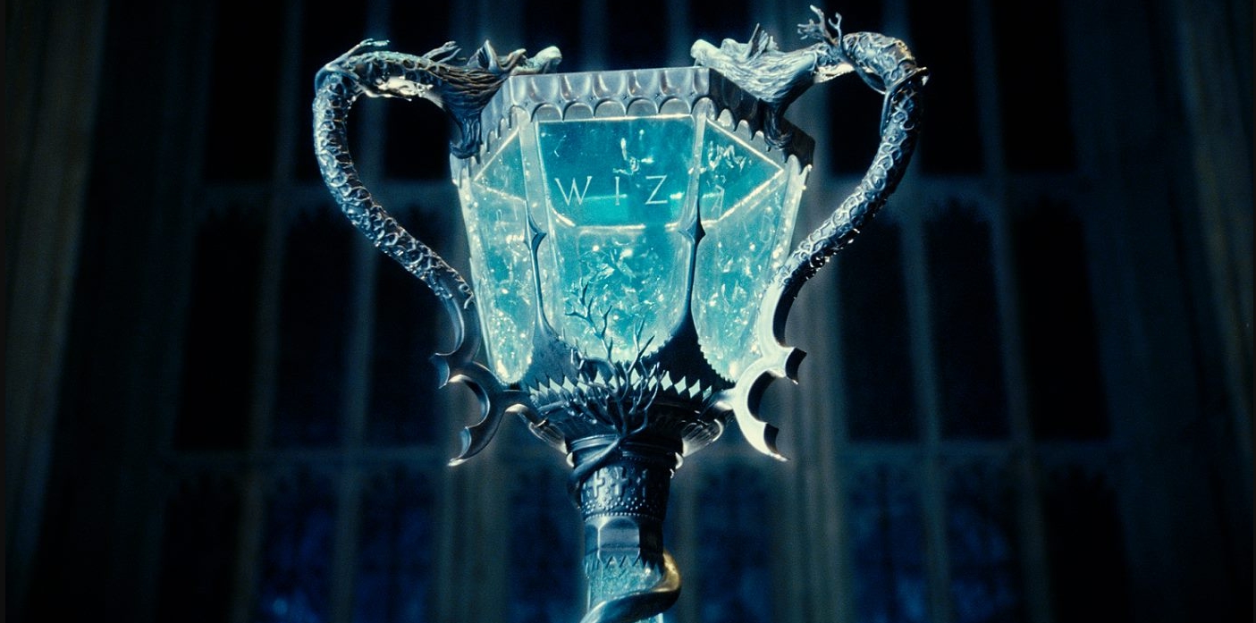 A imagem mostra a taça do torneio tribruxo, a qual é transparente com sua base e alças em um prata decorado. Dentro da taça há um líquido azul. Ao fundo há uma grande janela.