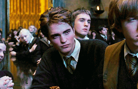 O gif mostra Robert Pattinson caracterizado como Cedrico Diggory. Ele usa um cardigan preto por cima de uma blusa social branca e uma gravata listrada em preto e amarelo. Ele está sorrindo e sendo parabenizado por seus amigos, os quais aplaudem. 