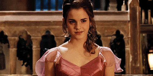 O gif mostra Emma Watson caracterizada como Hermione Granger descendo um lance de escadas. Ela está com o cabelo preso em um meio coque e usa um vestido cor de rosa em cetim, com mangas curtas em tule. Ela está sorrindo. 