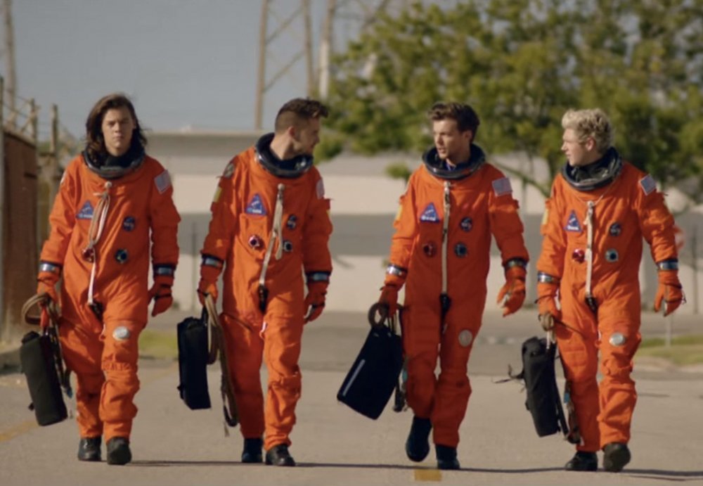 A imagem mostra os quatro integrantes do One Direction, vestindo roupa de astronauta, em cor laranja. Eles não usam capacete e estão andando em direção à câmera.