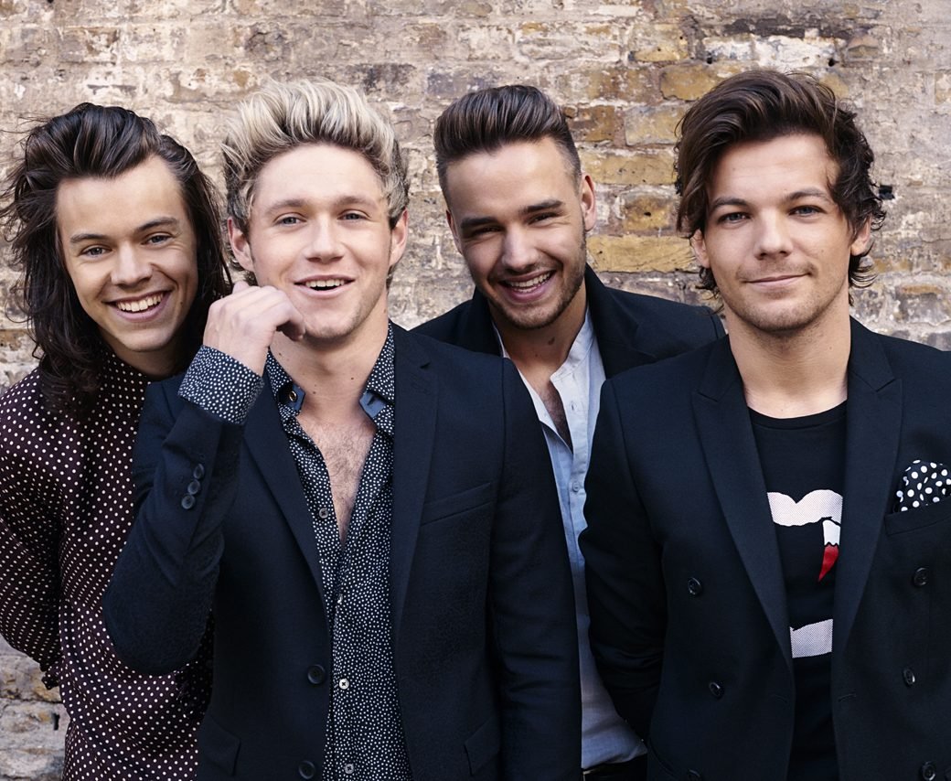 Na imagem, os quatro integrantes da banda One Direction olham para a câmera. Eles vestem traje social despojado. Harry, Niall e Liam estão sorrindo mostrando os dentes, Louis está sorrindo de boca fechada.