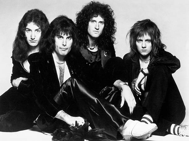 Imagem em preto e branco dos quatro integrantes da banda sentados no chão. Da esquerda para a direita estão John Deacon, Freddie Mercury, Brian May e Roger Taylor. Todos eles estão vestindo roupas pretas e possuem cabelos compridos.