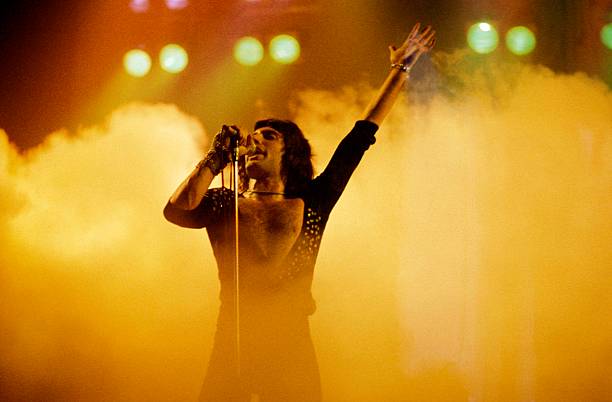 Imagem de Freddie Mercury no palco. Ele usa um macacão preto com decote em v até o meio da barriga, cabelos médios pretos e está segurando um microfone na mão direita. Sua mão esquerda está levantada para cima e ao fundo vemos uma fumaça alaranjada.
