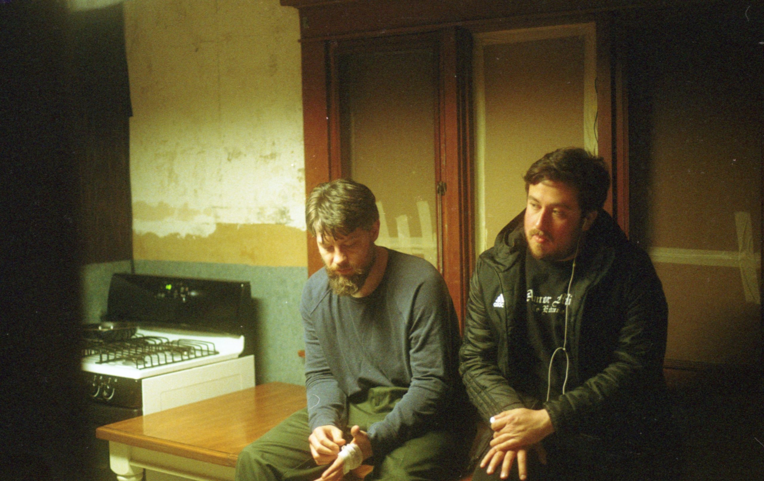 O ator Patrick e o diretor Jonathan, dois homens brancos, de barba e na casa dos trinta anos, estão sentado numa mesa, nenhum deles olha para a câmera.