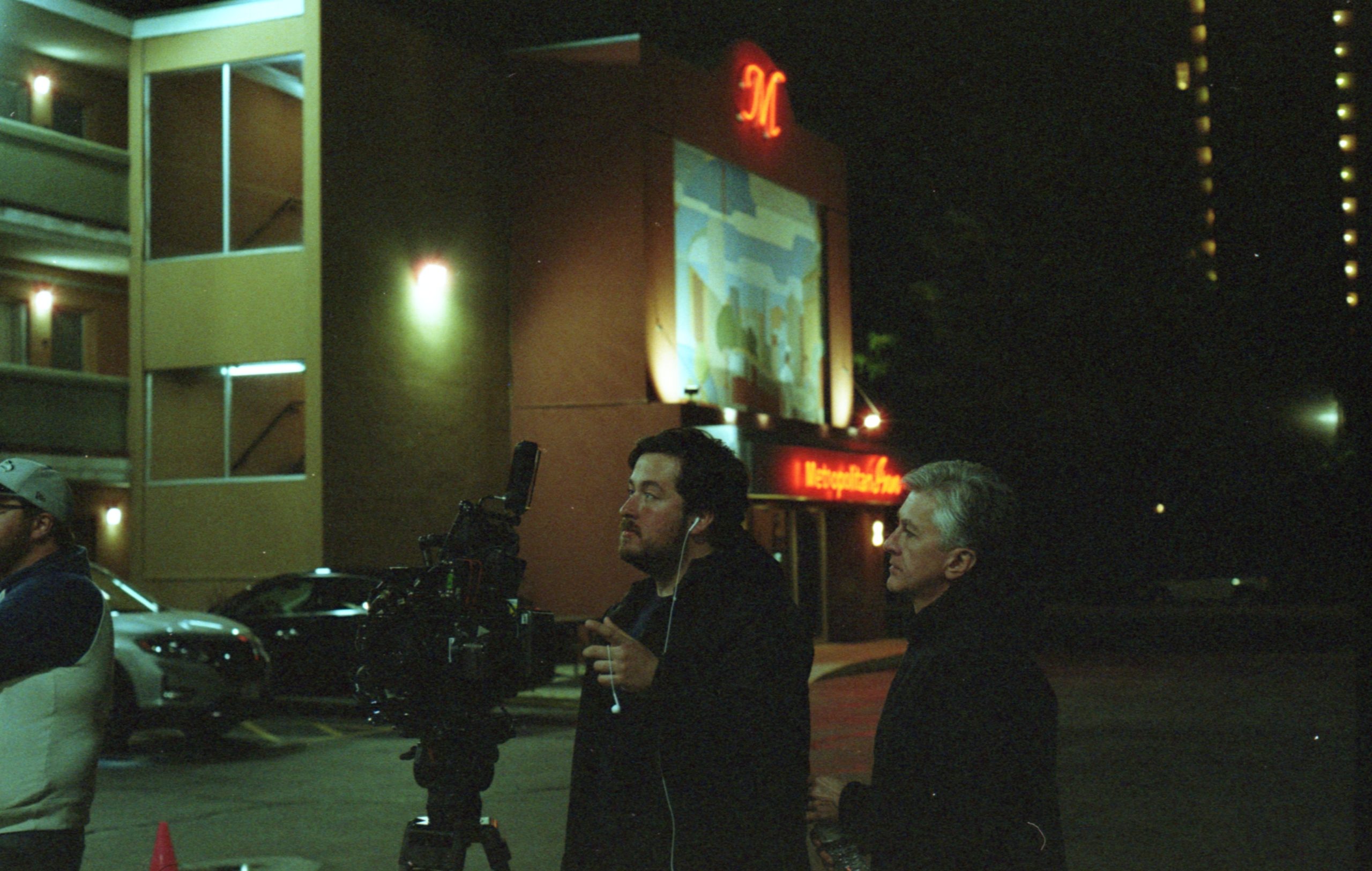 O diretor Jonathan segura a câmera, olhando para o que ela filma. Ele está ao lado de um homem mais velho e grisalho, eles parecem estar num estacionamento, com carros ao fundo e um visor vermelho na entrada de algum estabelecimento no fundo. 