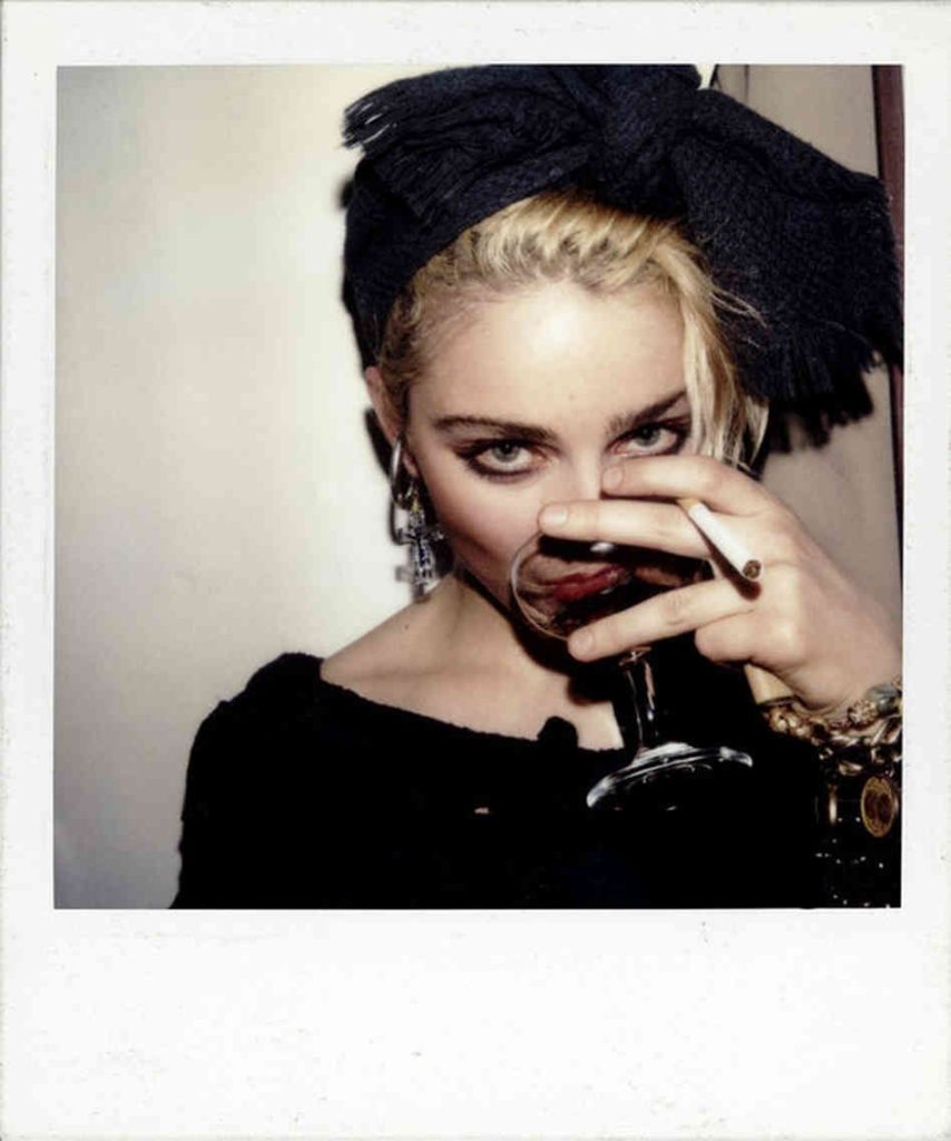 Foto em estilo Polaroid, com bordas brancas. Na parte da imagem, há a cantora Madonna quando ainda jovem adulta. Ela é uma mulher branca, de cabelos loiros, e usa um laço preto na cabeça. Ela bebe uma taça de vinho, com um cigarro entre os dedos e está olhando para a câmera.