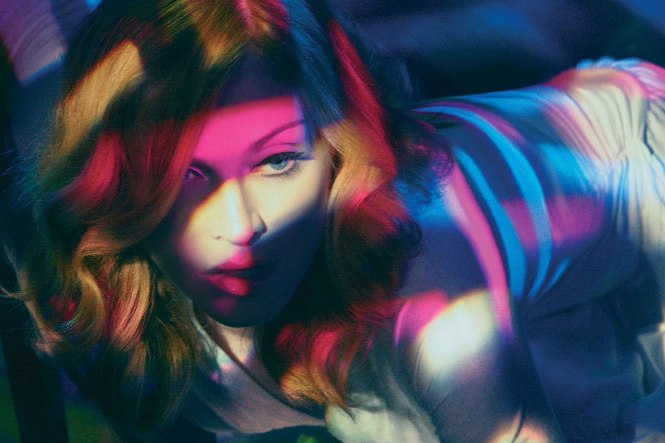 A foto mostra apenas a cabeça e o torso da cantora Madonna. Ela está apoiada de bruços, e está sendo iluminada por flashes de luz rosa, amarelo claro e azul. Ela não está olhando para a câmera.
