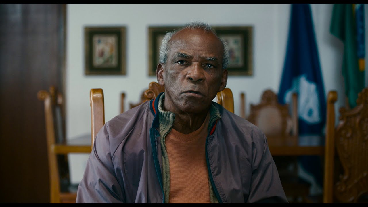 Na foto, vemos uma cena do filme Casa de Antiguidades. Antonio Pitanga interpreta m homem negro e idoso, que olha diretamente para a câmera, ele usa camiseta amarela e blusa cinza, ele está numa sala, com quadros e uma mesa atrás, desfocados.