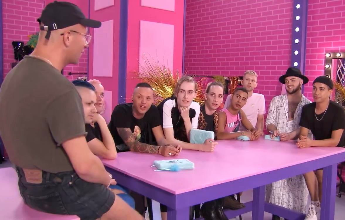 10 drag queens, todas desmontadas, estão ao redor de uma mesa rosa e roxo, os detalhes do cenário são da mesma cor