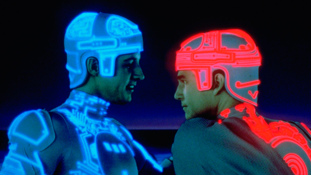 A imagem mostra dois homens brancos se encarando de perfil. O primeiro usa um capacete e uma roupa branca com fios neons azuis por toda a extensão. O segundo usa uma roupa igual, porém seus fios neons são vermelhos.