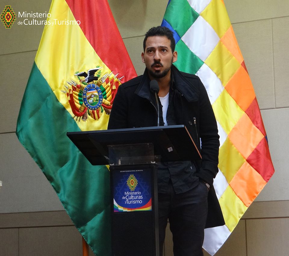 Bahman, um homem iraniano de 30 anos, está em frente à duas bandeiras, uma delas é da Bolívia. Ele fala num palanque com o microfone próximo a seu rosto. Ele tem cabelos pretos curtos e barba e bigode pretos, veste jeans e uma jaqueta preta