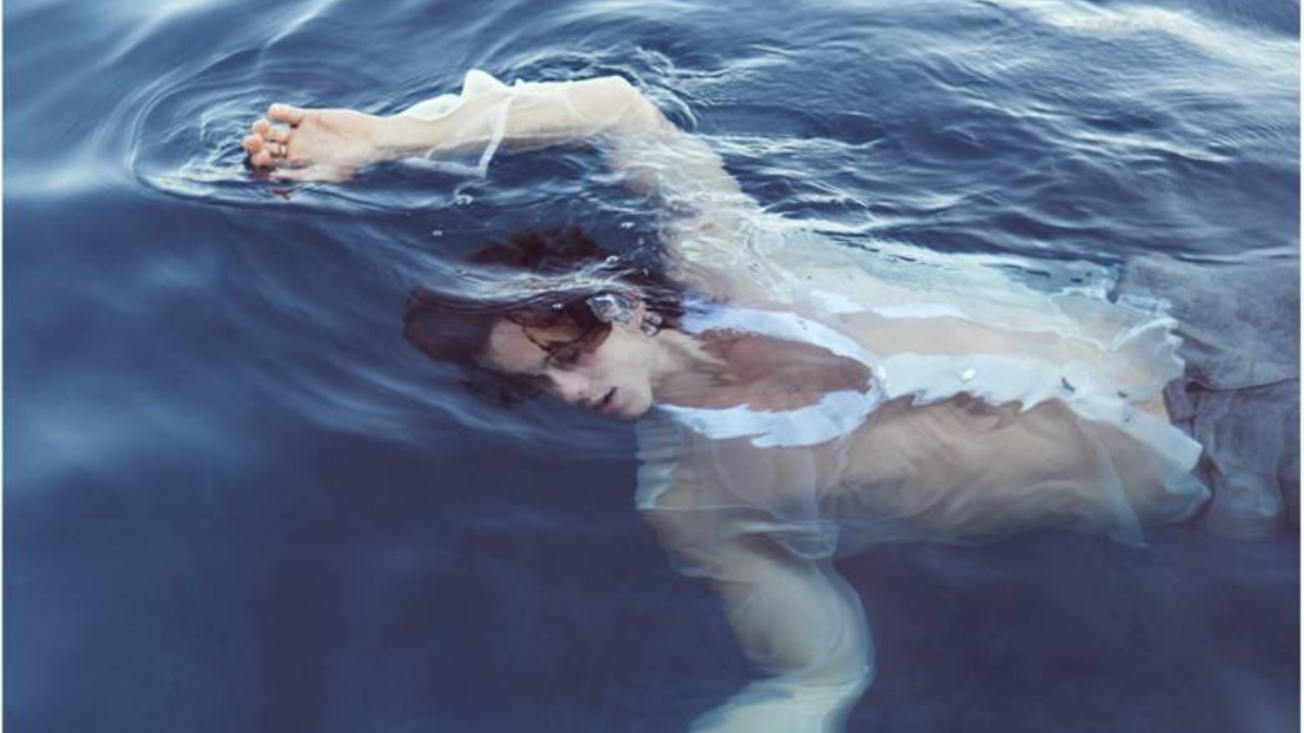 A imagem mostra Shawn Mendes submerso na água. Ele usa uma camisa branca que contrasta com o fundo azul da imagem.
