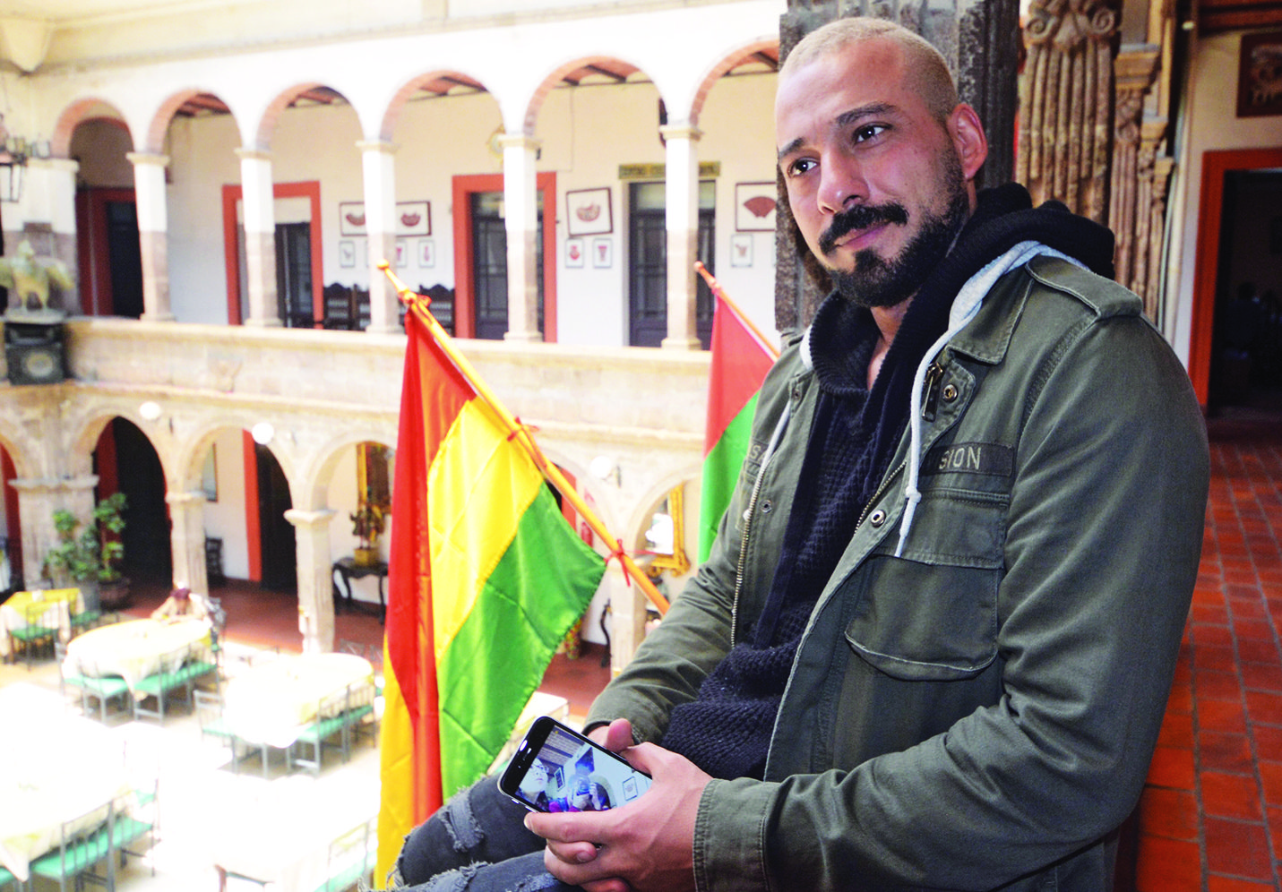 Bahman, careca e de barba e bigode preto, está sentado num banco próximo a duas bandeiras, ele usa jaqueta verde e segura o celular nas mãos, em cima do colo