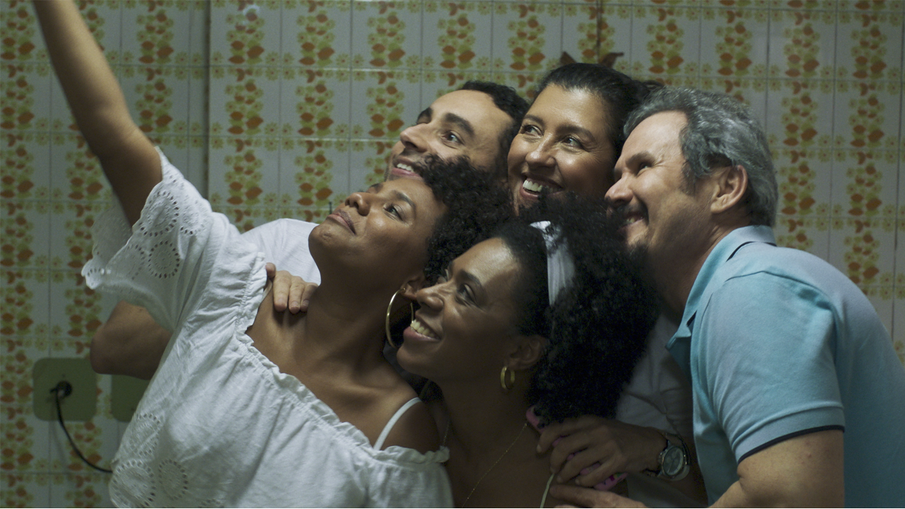 A imagem é uma das cenas do filme. Na imagem, Madá e os outros funcionários da casa estão posicionados em grupo sorrindo para uma selfie.