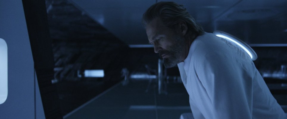 A imagem mostra o personagem de Kevin Flynn de perfil em uma sala branca escura. Ele tem cabelos e barba acinzentados e usa uma blusa branca larga. 
