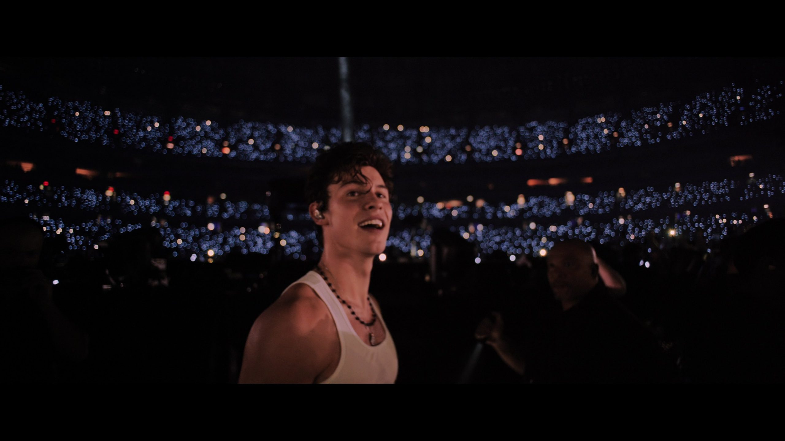 A imagem mostra Shawn Mendes com uma regata branca e cabelos curtos. Ele usa um colar de bolinhas pretas e está em frente a uma constelação de luzes.