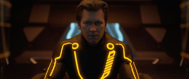 A imagem mostra o personagem Clu. Ele usa um macacão preto com fios neons laranja e tem o cabelo castanho claro e curto penteado para trás. Ele se parece com uma versão mais nova do personagem de Kevin Flynn. 