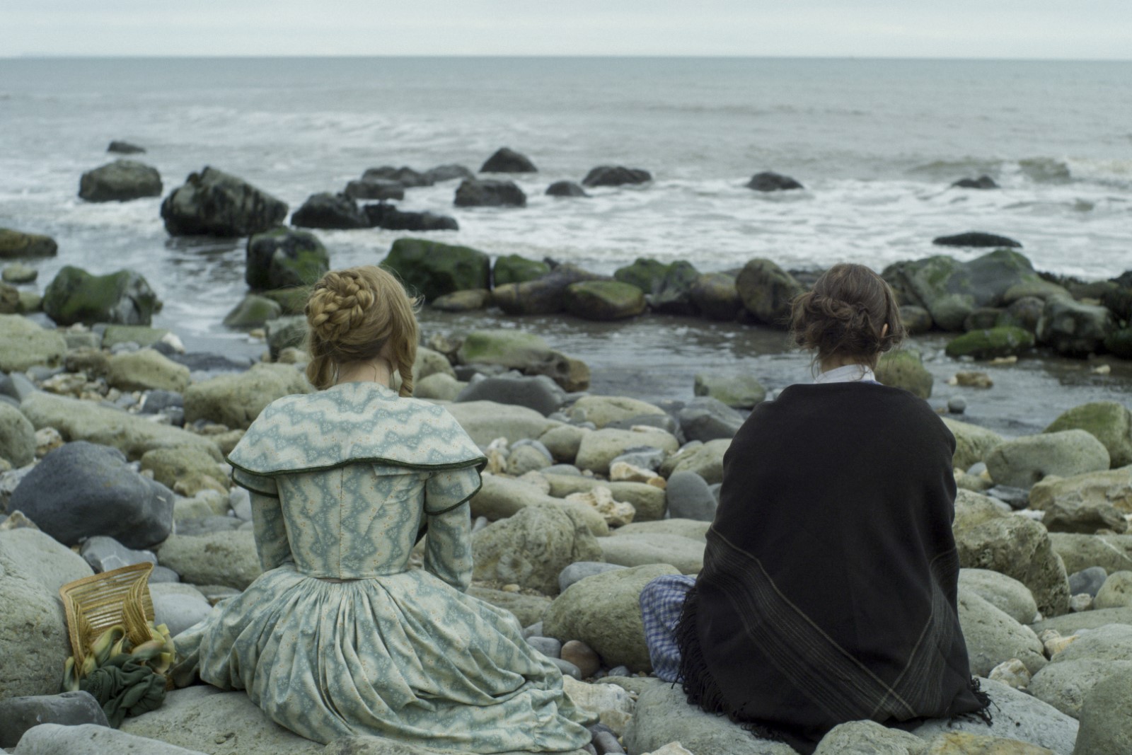 As duas mulheres estão sentadas nas pedras olhando para o mar. Saoirse, à esquerda, está usando um vestido azul claro e Kate, à direita, está de preto