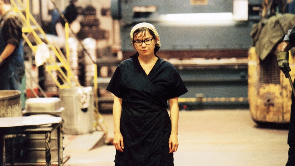 Cena do filme Dançando no Escuro. A imagem mostra Björk vestida de funcionária de fábrica, no meio de um corredor com aparência industrial. Ela veste um avental preto, touca branca e óculos.