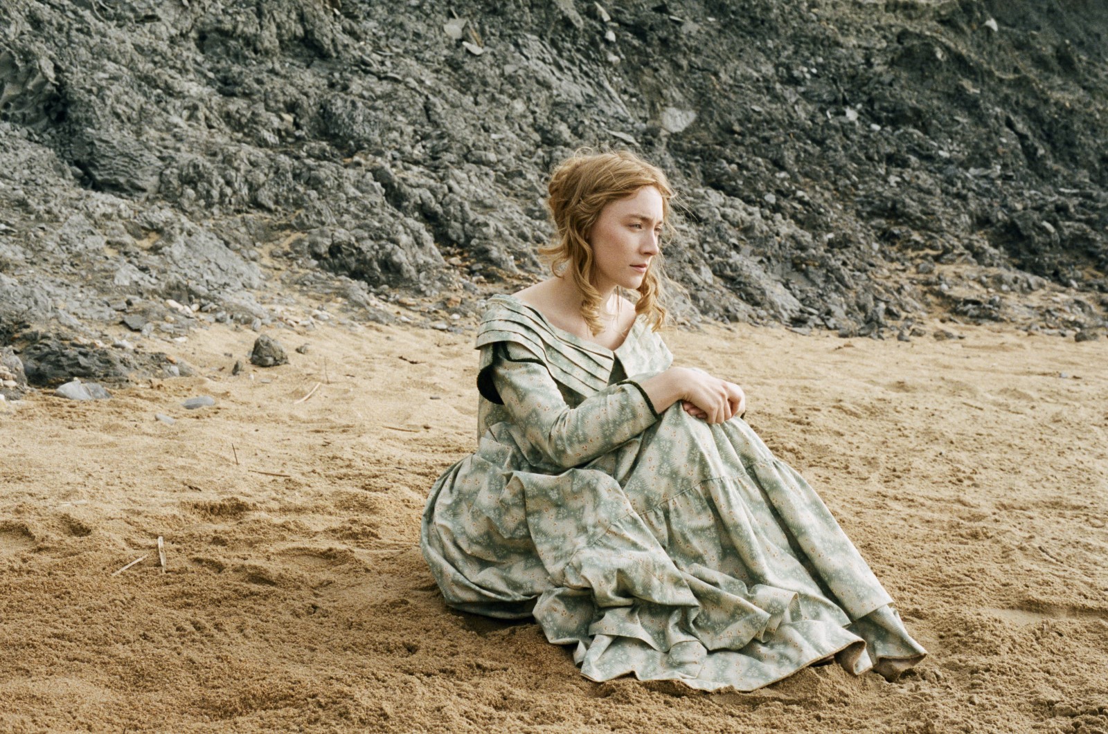 Saoirse está sentada sozinha na areia da praia. Ela tem os cabelos louros presos e usa um vestido com detalhes em verde. Atrás dela vemos uma grande rocha