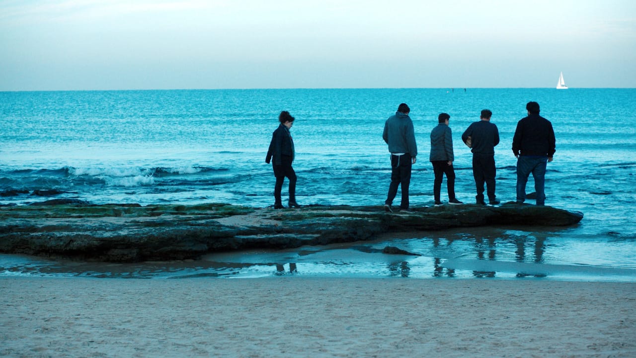5 pessoas estão reunidas às margens do oceano, uma delas está mais afastada à esquerda, o mar é azul e o céu é azul mais claro