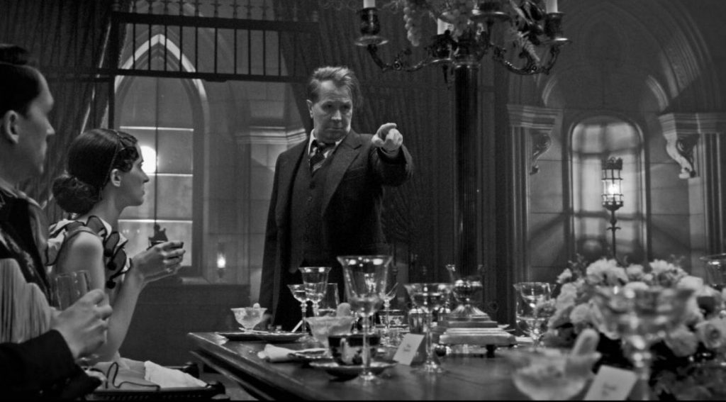 A foto está em preto e branco. Gary Oldman está em seu centro, usando um paletó e apontando o dedo esquerdo a frente. À esquerda, há duas pessoas o encarando. Ele está em frente a uma mesa, com diversos talheres, pratos e copos em cima. Acima dele, há um lustre.