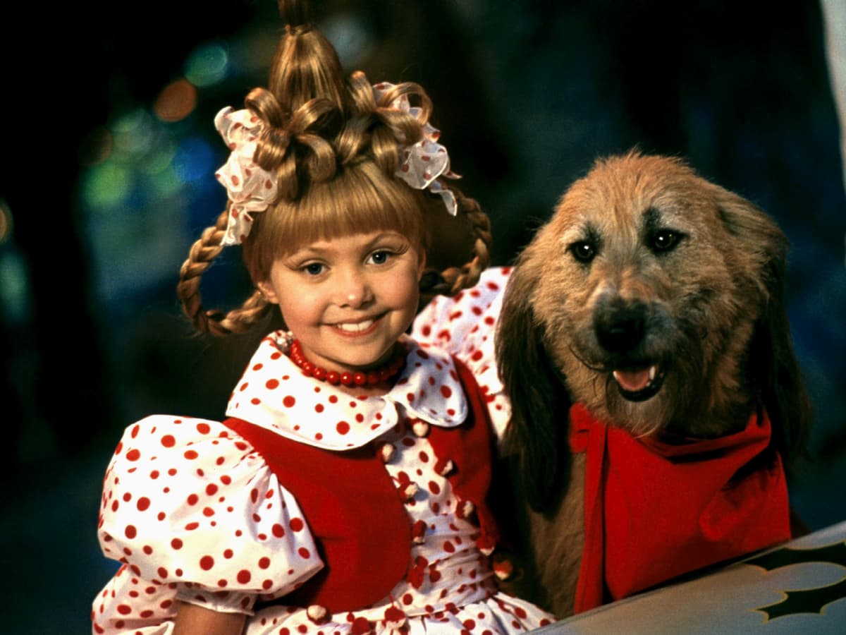 A imagem mostra a personagem Cindy Lou Quem ao lado do cachorro Max. Ela usa um vestido vermelho com mangas bufantes em branco com bolinhas vermelhas. Seu cabelo está preso para cima e é adornado com tranças. Max tem pelos castanhos claros e usa um cachecol vermelho. 