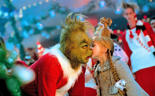 A imagem mostra o personagem Grinch vestido como um Papai-Noel, com um grande casaco vermelho. Ao seu lado Cindy Lou Quem veste um casaco bege e seu cabelo está preso em um coque alto com longas tranças soltas na lateral. Ela está beijando a bochecha de Grinch que parece surpreso.