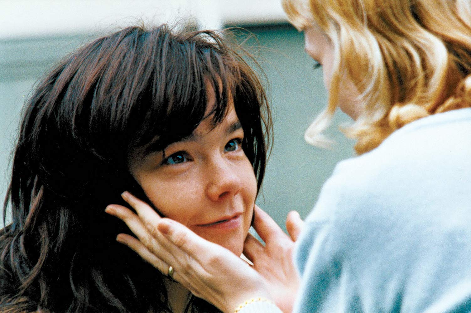 Cena do filme Dançando no Escuro. A imagem mostra uma mulher loira com as mãos no rosto de Björk, que olha para seu rosto e sorri. 