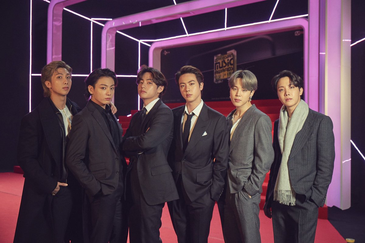 Foto do BTS no prêmio Mnet Asian Music Awards. Todos os membros do grupo estão usando terno e olham diretamente para a câmera com expressões sérias, ou com as mãos nos bolsos das calças ou com os braços cruzados. Ao fundo, atrás deles, está o palco do prêmio, com um chão vermelho e fios de luzes brancas.