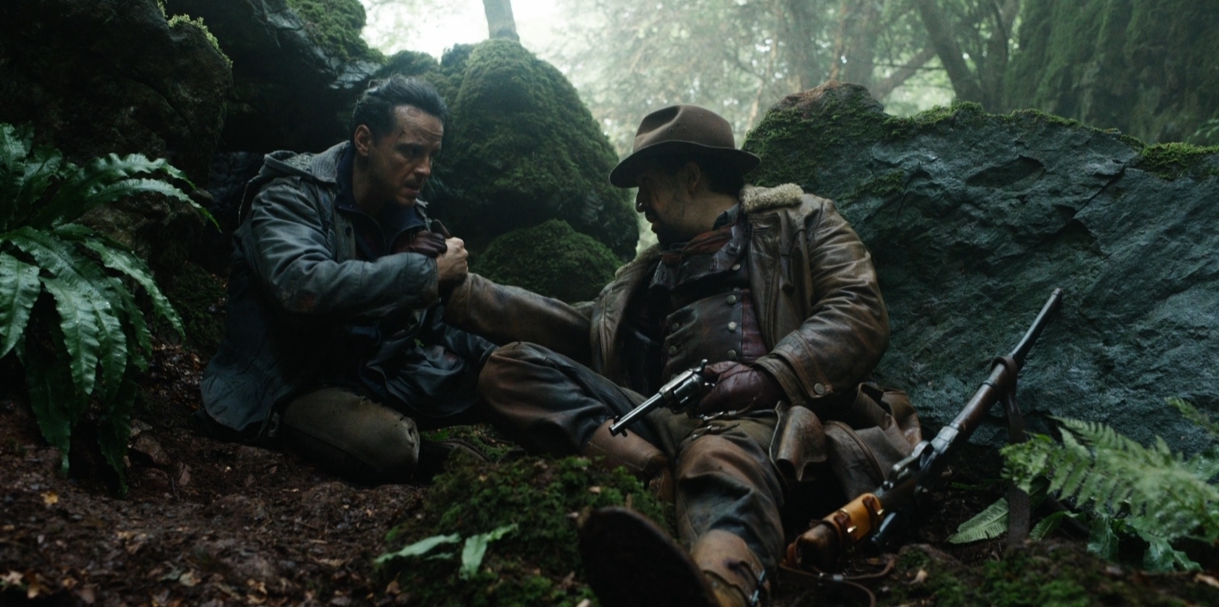 Na imagem, John Parry e Lee Scoresby estão sentados na floresta. Eles estão apertando as mãos e olham um para o rosto do outro. A aparência é de cansaço, e Lee segura uma arma na mão.