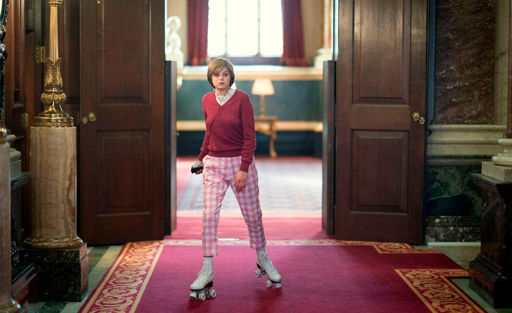 Na imagem, Lady Di anda de patins pelo palacio real, a personagem utiliza uma calça xadrez rosa claro e um sueter vinho.