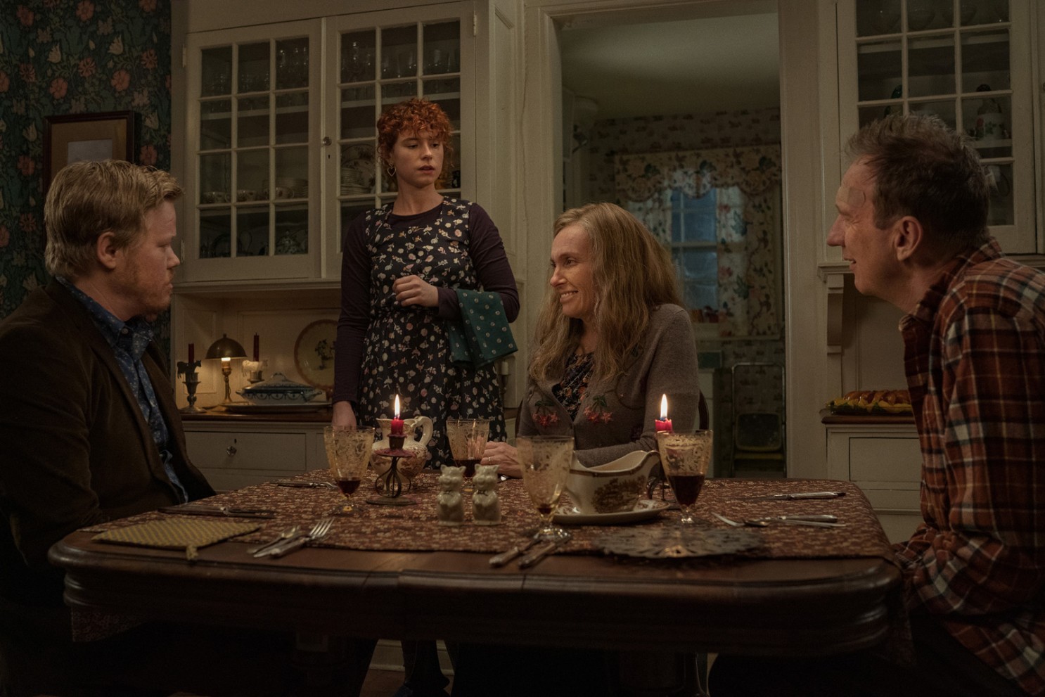 Três adultos estão sentados à mesa, enquanto a jovem mulher está de pé. O ambiente é mal iluminado e tem tons terrosos
