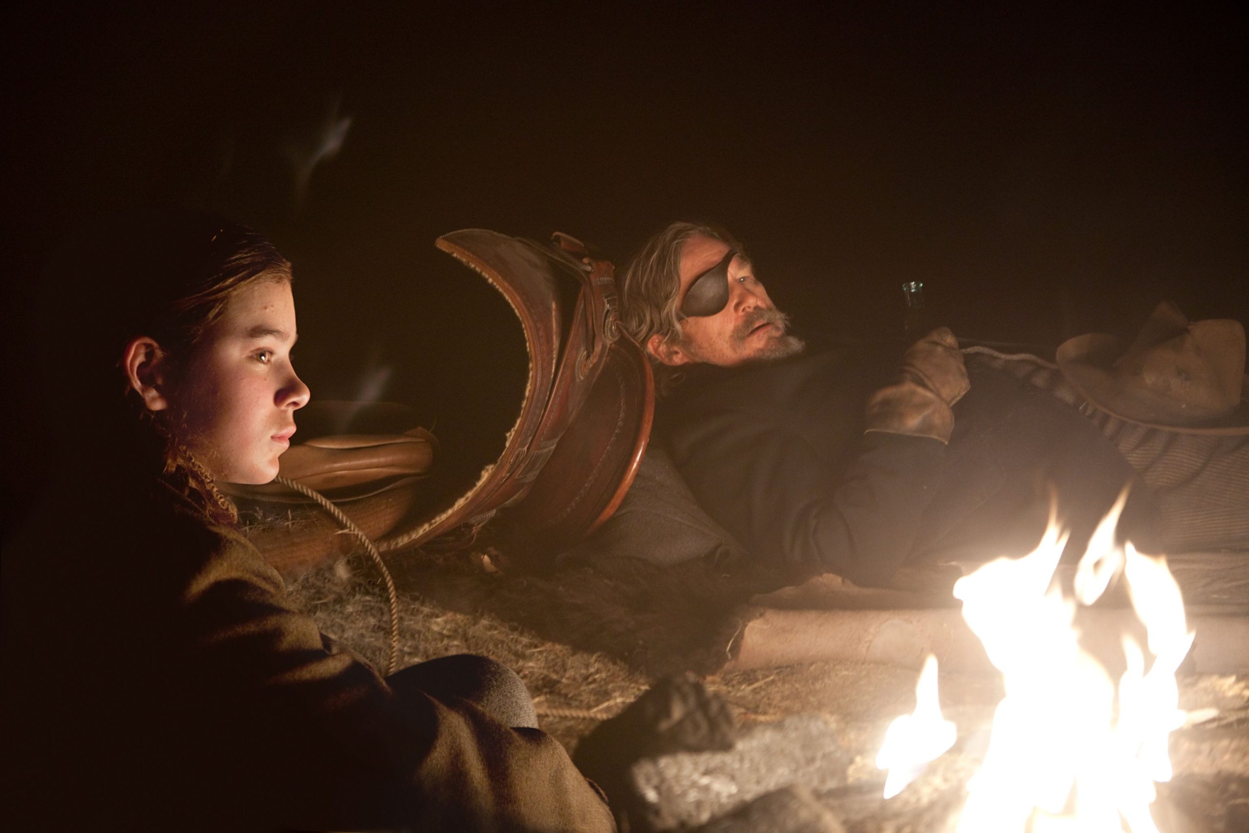A imagem mostra uma garotinha e um homem usando tapa-olho em volta de uma fogueira. Eles estão olhando ambos para a frente. O homem está deitado. É noite. 