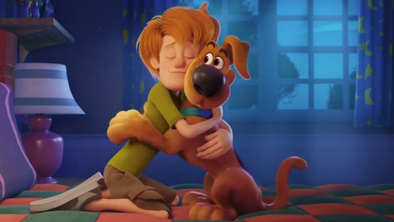 A imagem mostra Salsicha e Scooby abraçados em cima de uma cama. Eles usam seus figurinos clássicos, sendo o de salsicha a blusa verde, bermuda cáqui e tênis e de Scooby a coleira azul. 