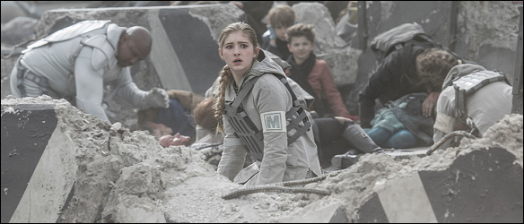 A imagem mostra Prim em meio aos destroços como uma trança loira lateral. Ela usa uma roupa militar cinza e ao fundo vemos pessoas machucadas.