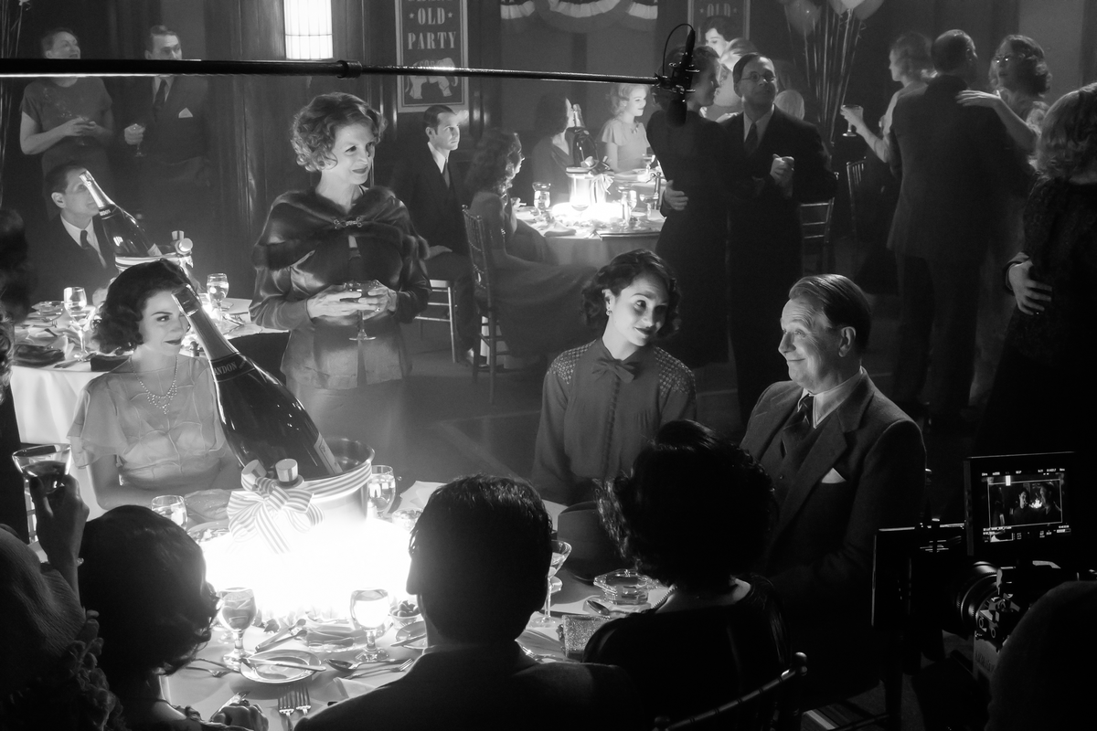 A imagem está em preto e branco. Vemos uma mesa com um balde com champagne dentro. Ao redor dela, três mulheres olham para a figura de Gary Oldman, que sorri. De fundo, temos outras mesas e casais dançando. Ao redor, vemos um microfone e uma câmera de filmagem, indicando os bastidores.