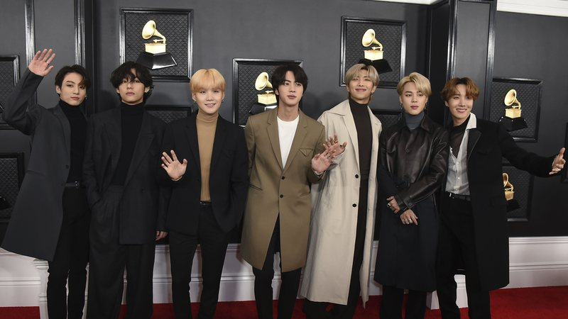 Foto dos membros do BTS no tapete vermelho do Grammy em 2020. O grupo é composto por sete jovens coreanos. Quatro deles acenam para as câmeras e todos tem os cabelos lisos na altura da orelha. Atrás deles está o painel do Grammy, de fundo preto com quadros do gramofone que é a figura do prêmio.