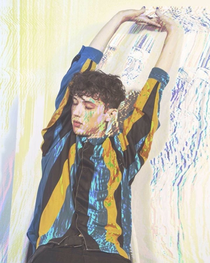A imagem mostra Troye Sivan se espreguiçando, de olhos fechados. Ele usa uma camiseta com listras verticais azuis, amarelas e pretas. Foi adicionado um efeito de glitch na imagem. 