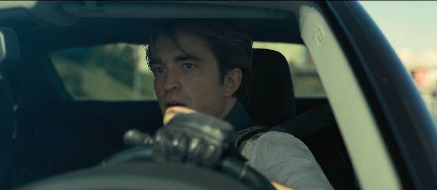 A cena mostra Robert Pattinson dirigindo um carro, com um mão no volante. Pattinson é um homem branco de 30 anos, ele tem cabelo castanho liso e um pouco longo, ele usa uma luva preta de couro na mão esquerda.