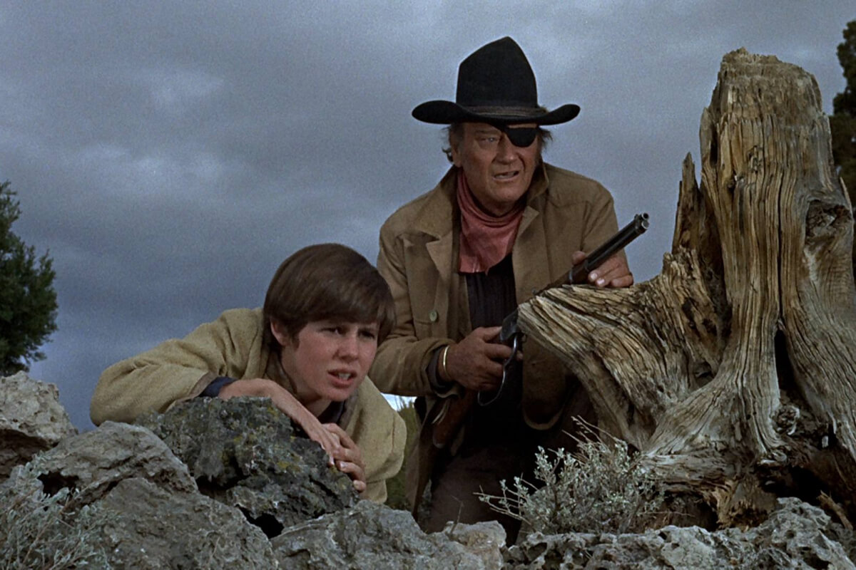 Cena do filme Bravura Indômita de 1969. A imagem mostra um garotinho e um homem se escondendo atrás de uma rocha. O homem usa tapa-olho e chapéu, e tem uma arma na mão.