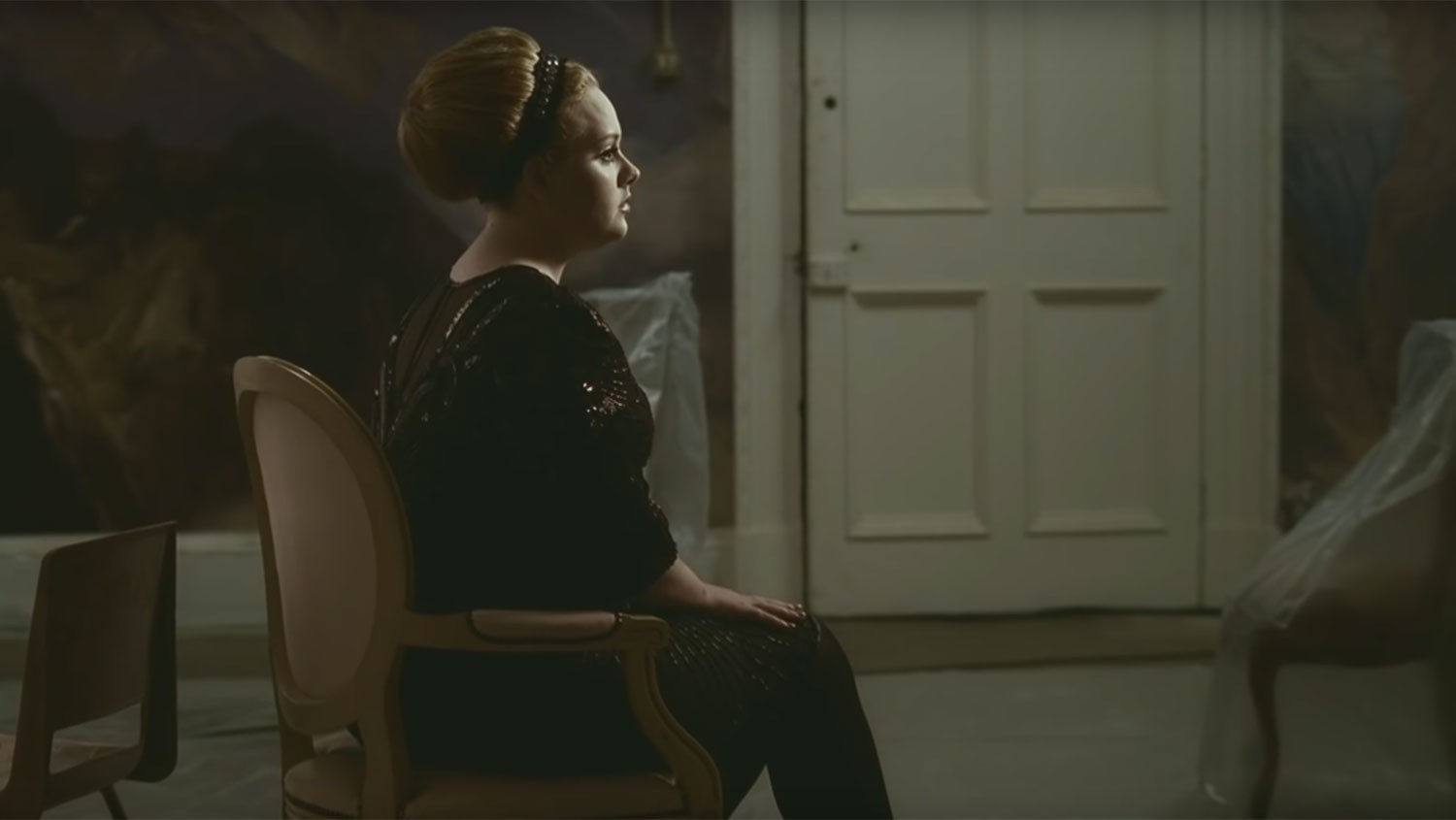 Imagem do clipe Rolling In The Deep. Mostra Adele sentada de perfil em uma cadeira grande. Ela tem os cabelos loiros presos em um coque e usa um vestido de manga três quartos e meia calça, ambos pretos. Ao fundo há uma grande porta branca.