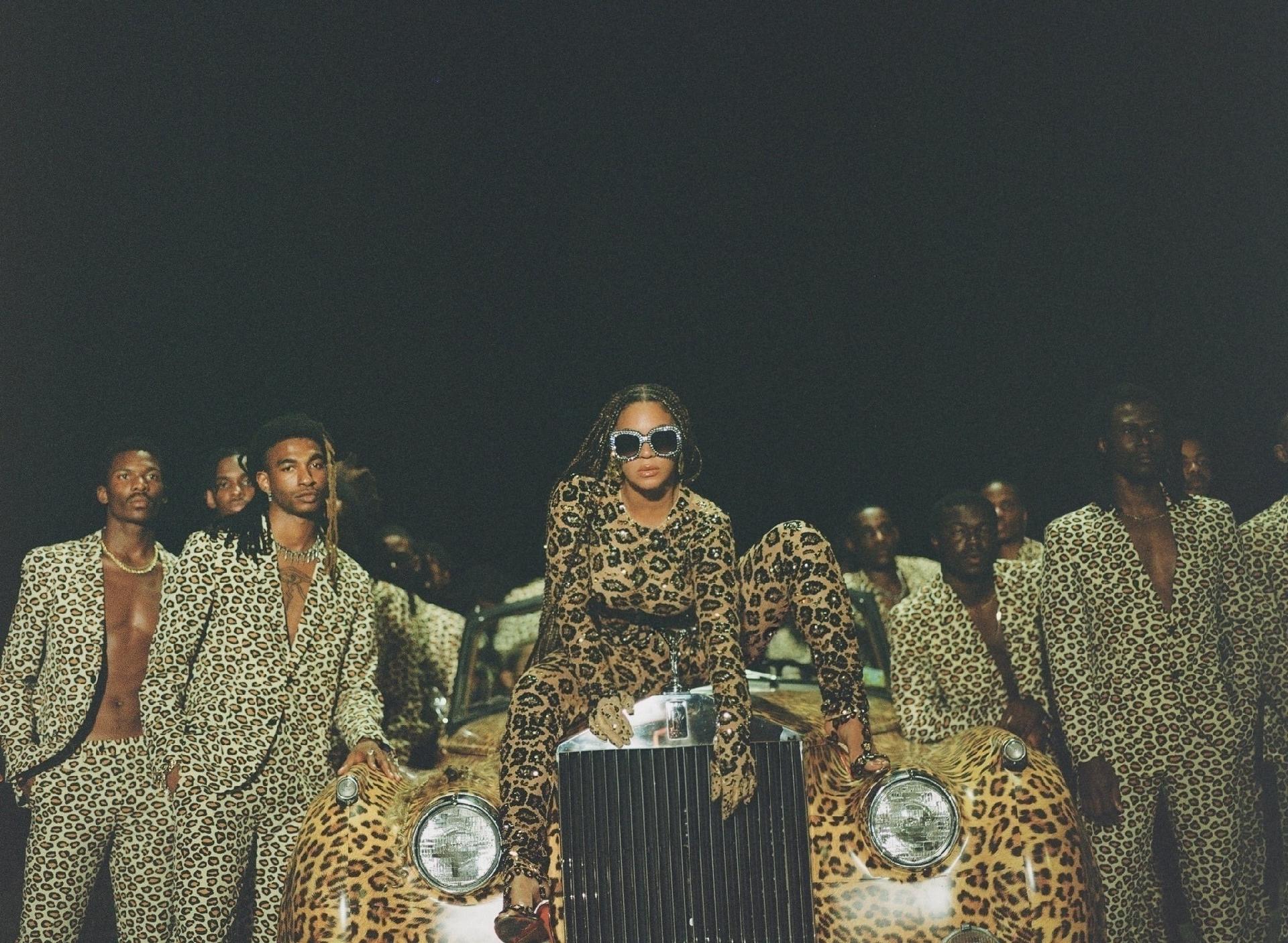 A imagem é uma foto do filme Black Is King. Nela, a cantora Beyoncé está sentada em cima de um carro com estampa de oncinha. Beyoncé é uma mulher negra, com cabelos longos em tranças, ela veste uma calça, blusa de manga comprida e um salto de estampa de oncinha e usa um óculos escuro no rosto. Ao redor do carro, estão vários homens negros com terno e calça de estampa de oncinha.