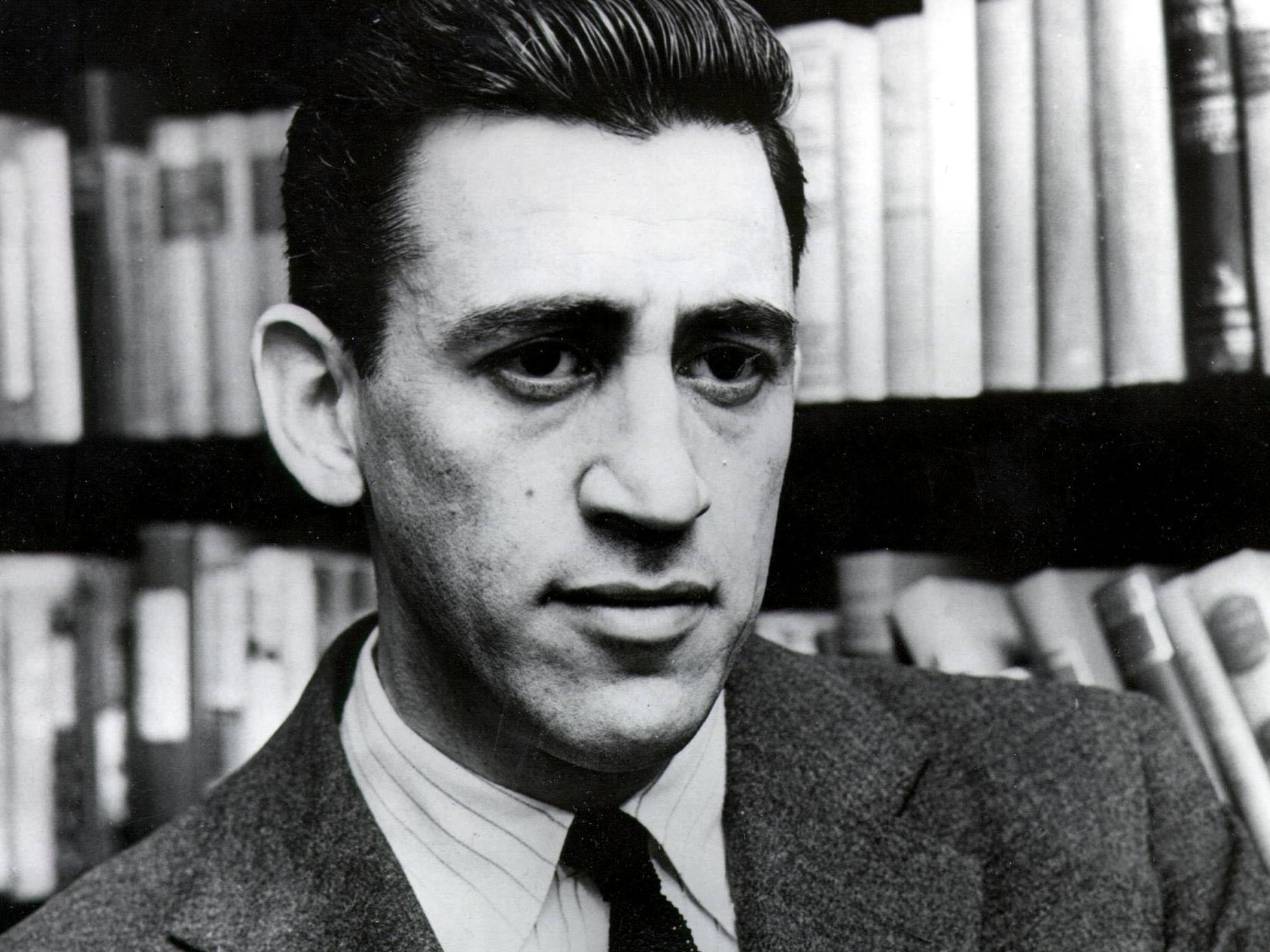 A imagem é uma fotografia em preto e branco do autor J.D. Salinger. Salinger era um homem branco de cabelos curtos lisos, formando um pequeno topete. Ele está vestindo um terno com uma camisa e gravata. Atrás dele, é possível ver uma estante de livros.