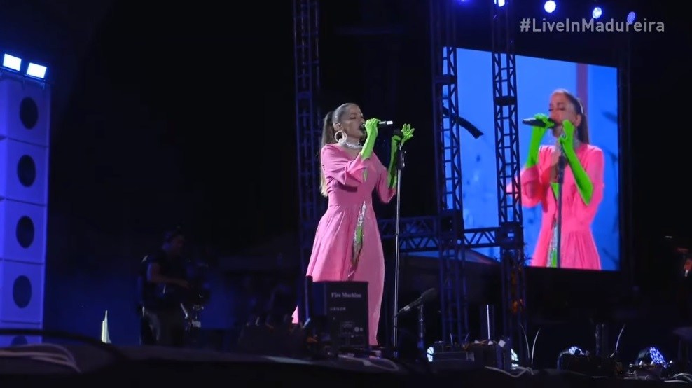 cena do show de Anitta no Parque de Madureira. Ela é branca, está no palco vestindo um robe rosa pink e usa luvas verde-limão que se estendem até seus cotovelos. Na mão direita ela segura o microfone enquanto canta para a plateia, e com a mão esquerda segura o pedestal do microfone.