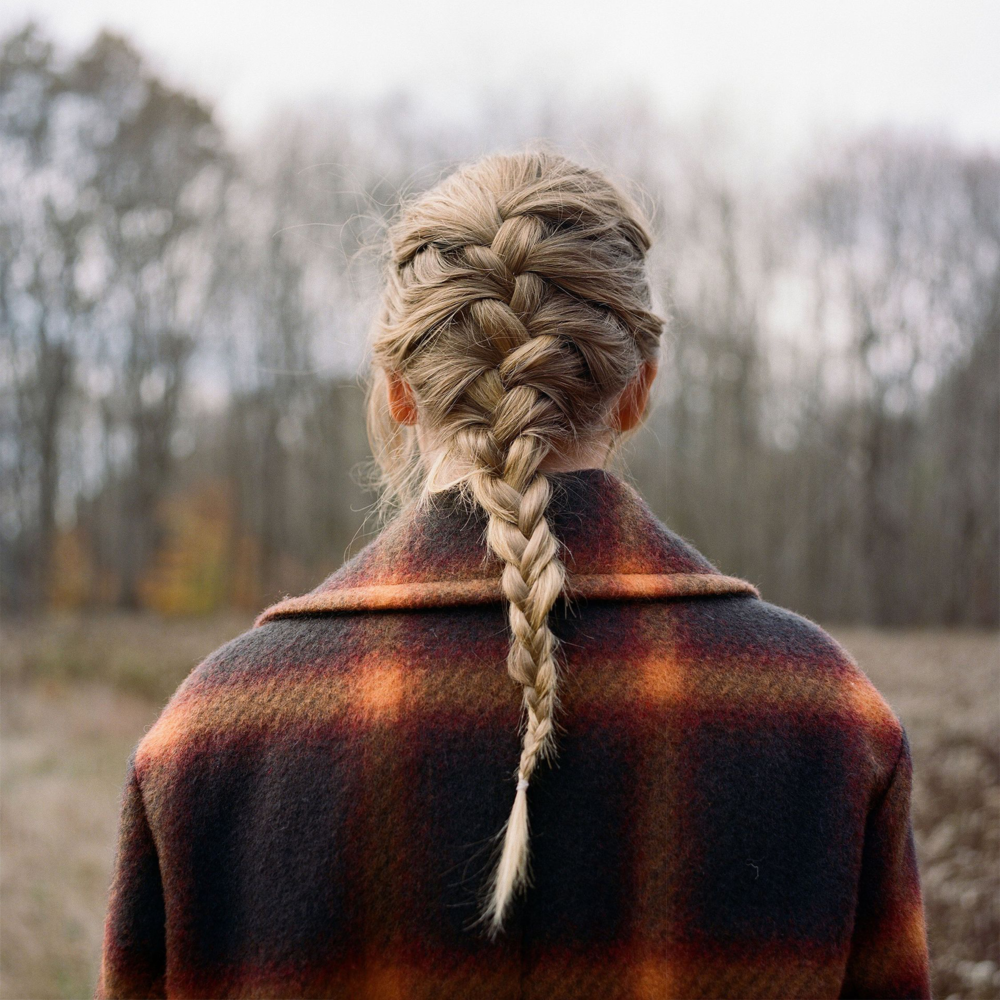 Capa do disco Evermore Deluxe Version. Taylor se encontra de costas, seu cabelo preso em uma trança, no meio de um campo gramado onde se pode ver árvores no fundo. 
