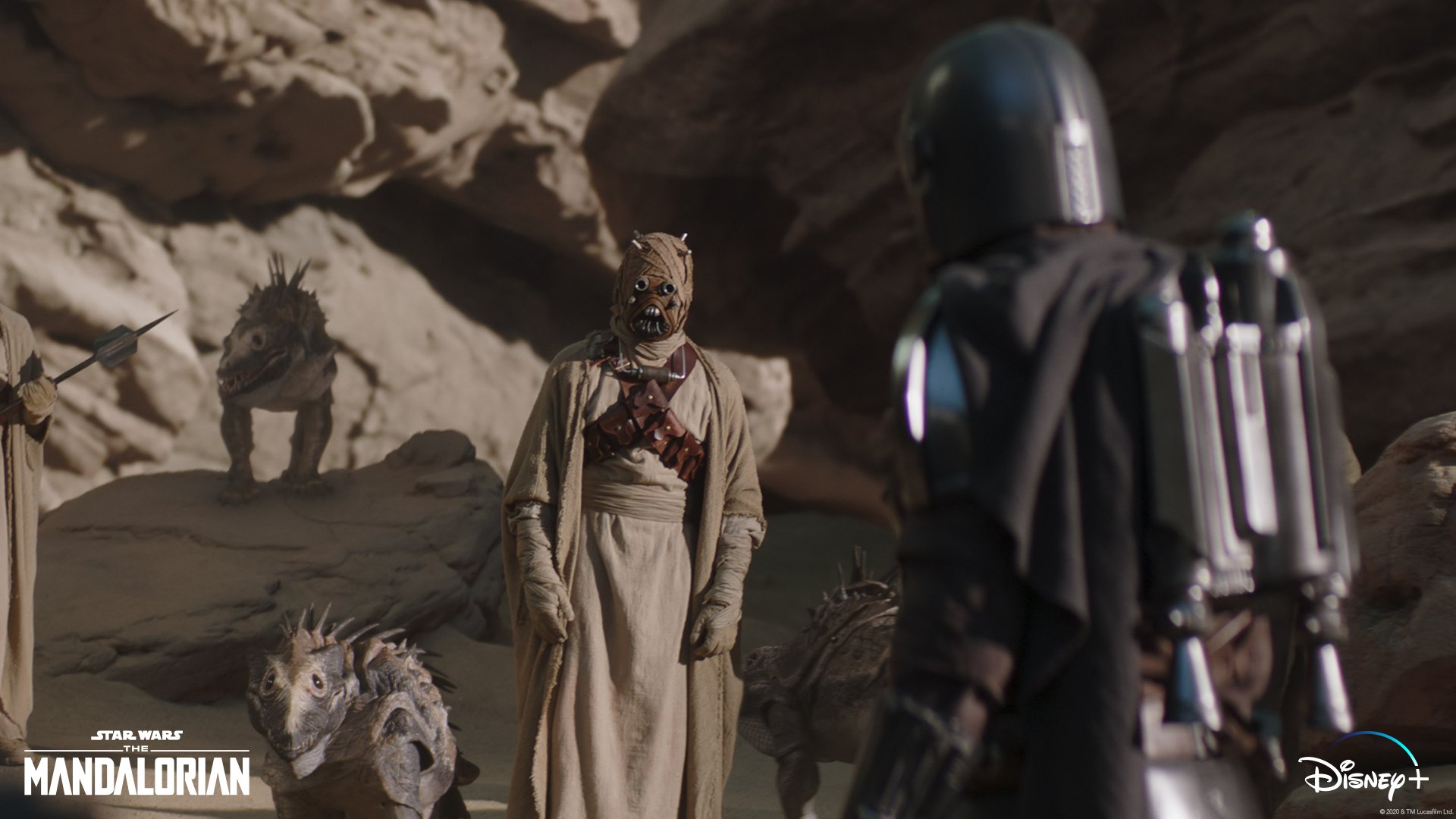 Cena da série The Mandalorian, em que Din Djarin, de costas à direita, conversa com uma criatura do Povo da Areia, que o encara, ao centro. Os dois estão próximos a pedras e há dois animais típicos do Povo, à esquerda.