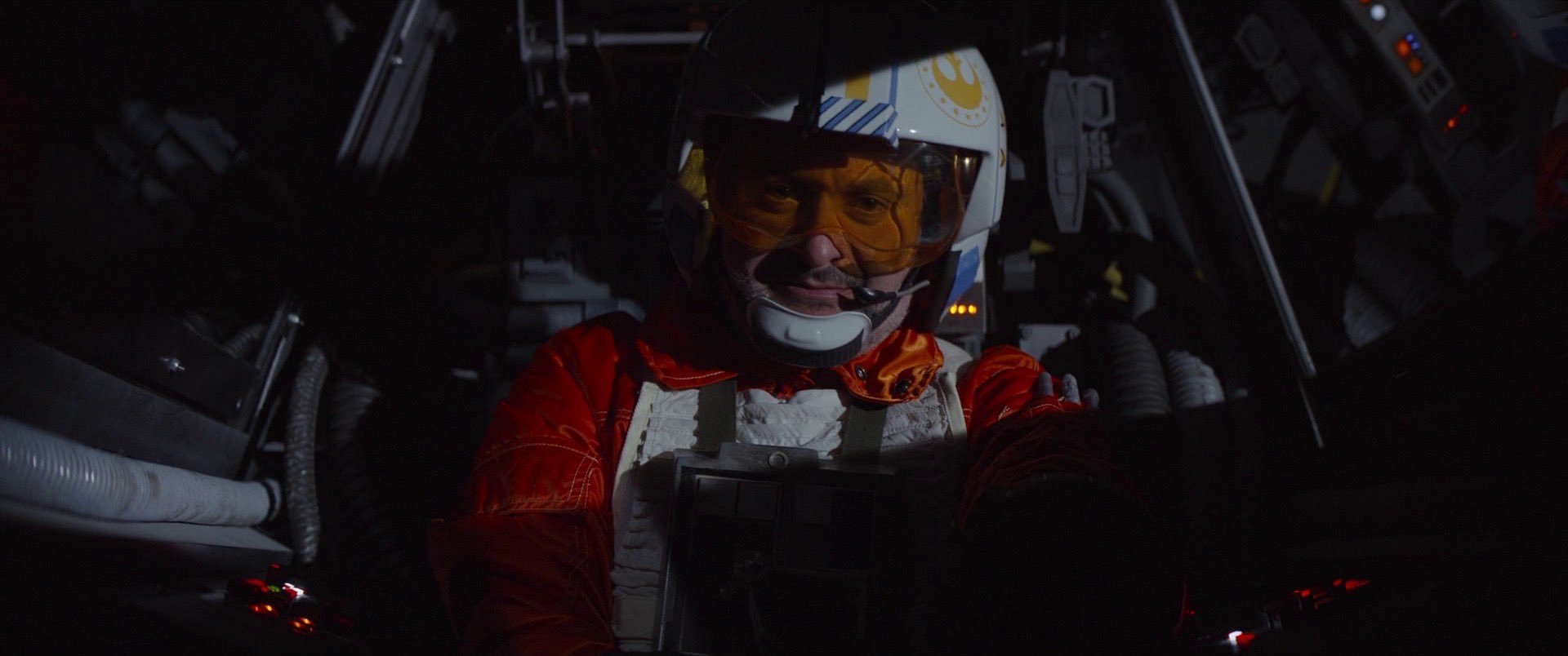 Cena da série The Mandalorian. Na imagem, há um piloto, interpretado por Dave Filoni, homem branco, vestindo um uniforme laranja e um capacete com o símbolo da Nova República. Ele está dentro de uma cabine da nave X-Wing.