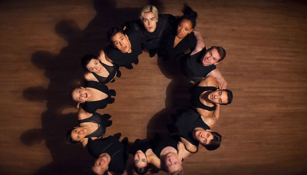 A imagem é de uma cena da série O Preço da Perfeição. A imagem é de 12 bailarinos se abraçando e olhando para cima. Todos eles vestem roupa preta e formam um círculo com seu abraço. 
